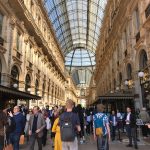 Pessoas circulam pelo interior da Galleria Vittorio Emanuele II, em Milão, sob a cobertura de vidro e a arquitetura histórica do local.
