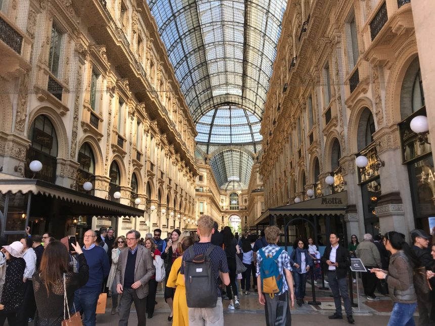 Pessoas circulam pelo interior da Galleria Vittorio Emanuele II, em Milão, sob a cobertura de vidro e a arquitetura histórica do local.