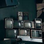 Pilhas de televisores CRT antigos em uma sala moderna com iluminação baixa e uma obra abstrata ao fundo