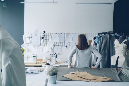 Designer de moda trabalha em um estúdio cercada por moldes de costura e peças de roupa.