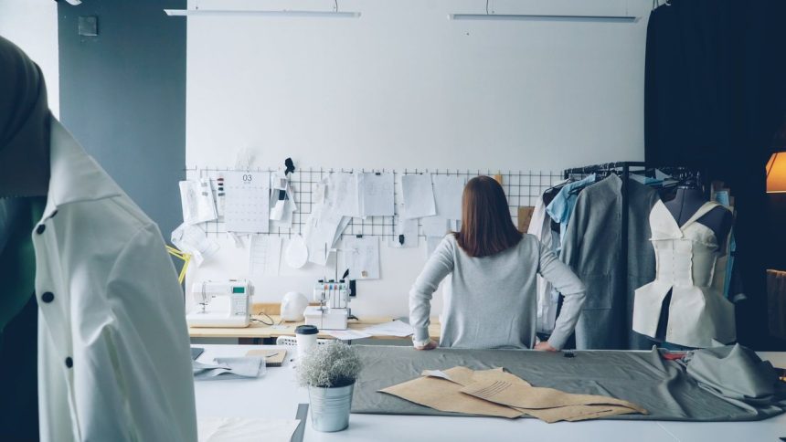 Designer de moda trabalha em um estúdio cercada por moldes de costura e peças de roupa.