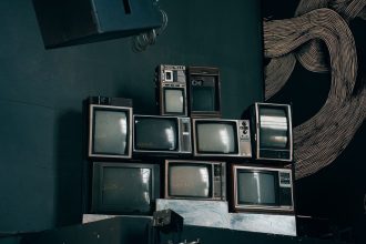 Pilhas de televisores CRT antigos em uma sala moderna com iluminação baixa e uma obra abstrata ao fundo